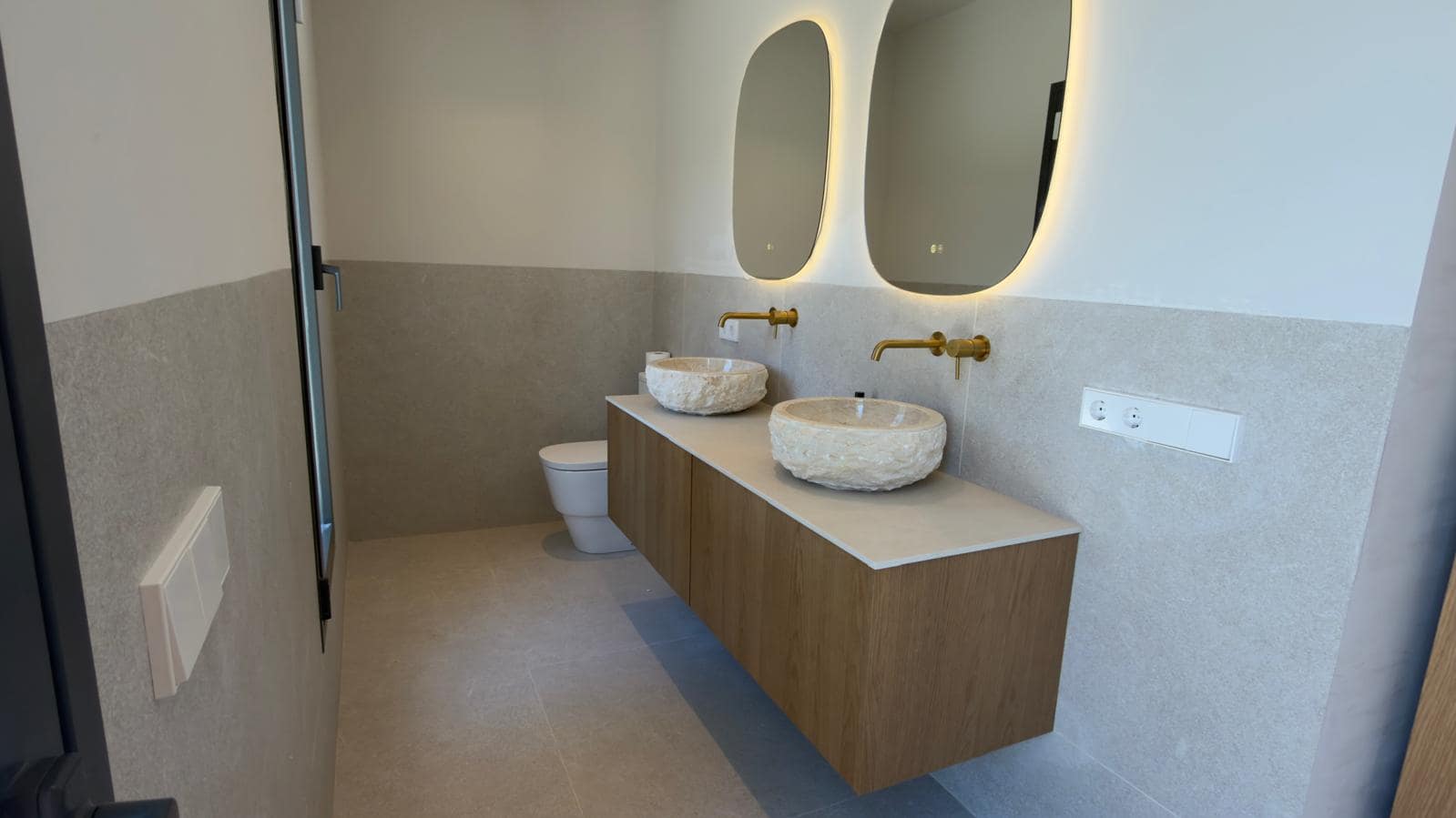 Baño reformado llave en mano en Mallorca con doble lavabo de piedra natural, grifería dorada y espejos ovalados retroiluminados